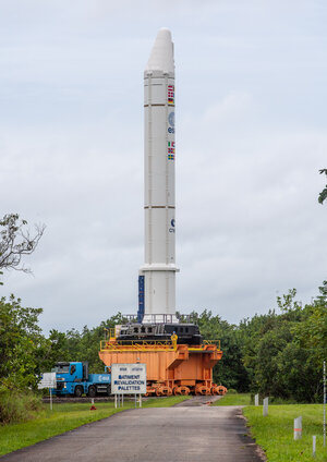 Launcher booster on top of a platform running along rails, pulled by a lorry. The image is taken outdoors, the sky is cloudy, in the foreground is a path and some trees